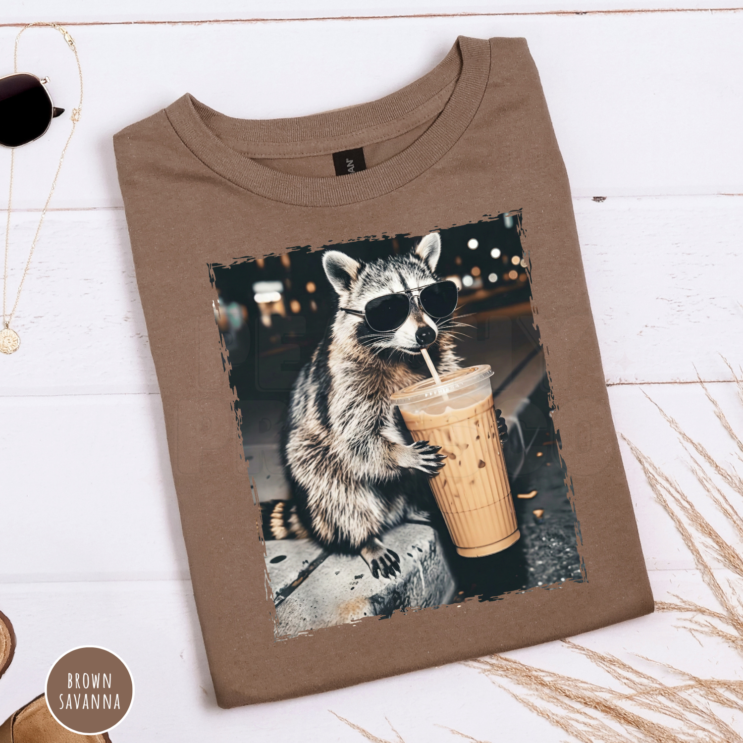 Brown Savanna Gildan graphic t-shirt folded flat featuring a funny raccoon wearing sunglasses while drinking an iced coffee. Cute coffee lover graphic tee with a vintage animal design.