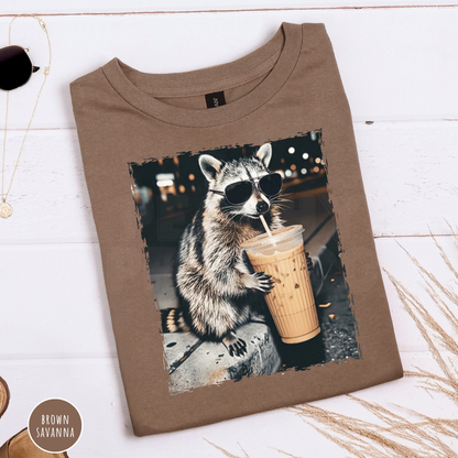 Brown Savanna Gildan graphic t-shirt folded flat featuring a funny raccoon wearing sunglasses while drinking an iced coffee. Cute coffee lover graphic tee with a vintage animal design.