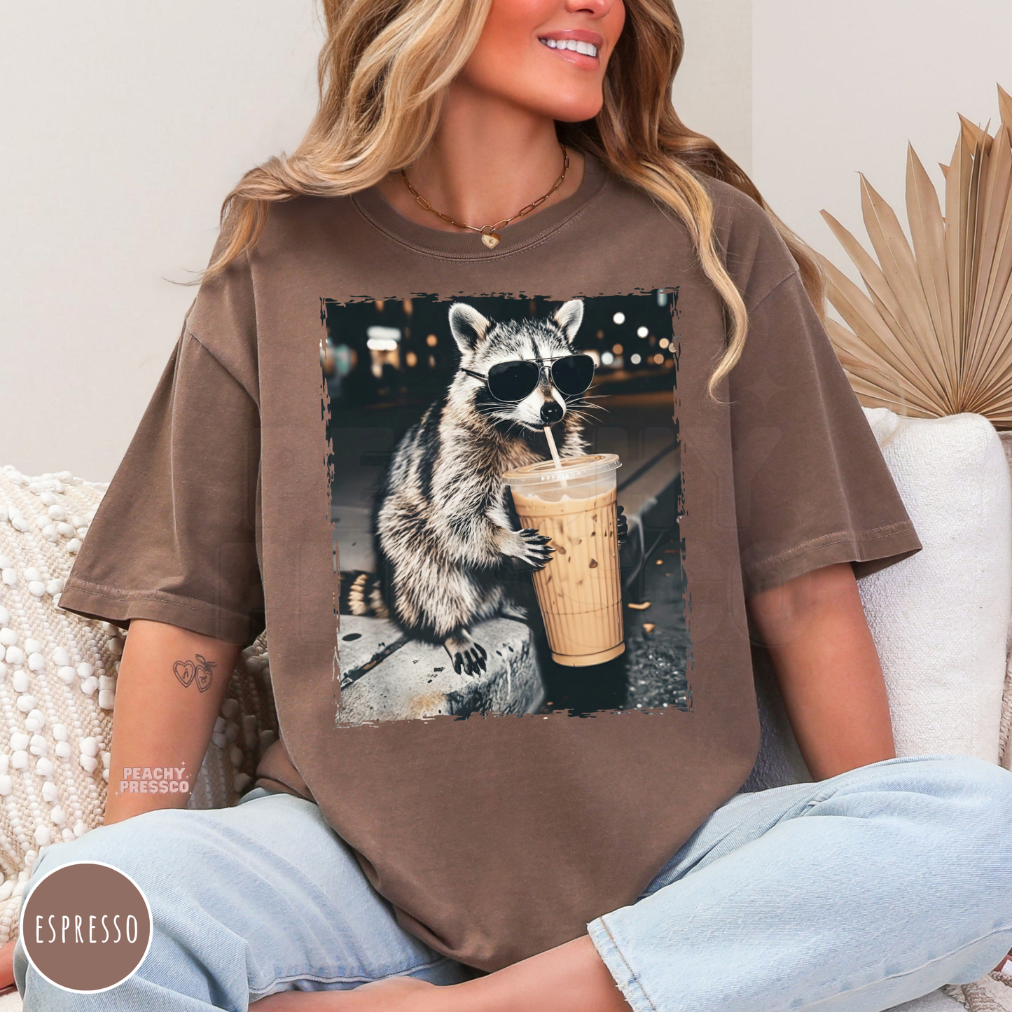 Woman wearing an espresso Comfort Colors graphic t-shirt featuring a cool raccoon wearing sunglasses while sipping an iced coffee. Vintage oversized coffee lover tee with a fun animal design.