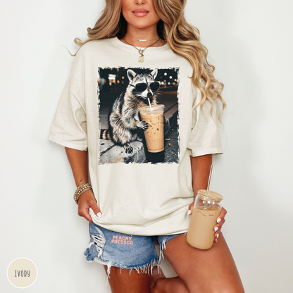 Woman wearing an ivory Comfort Colors graphic t-shirt featuring a funny raccoon wearing sunglasses while drinking an iced coffee. Cute oversized coffee lover tee with vintage aesthetic for caffeine lovers.