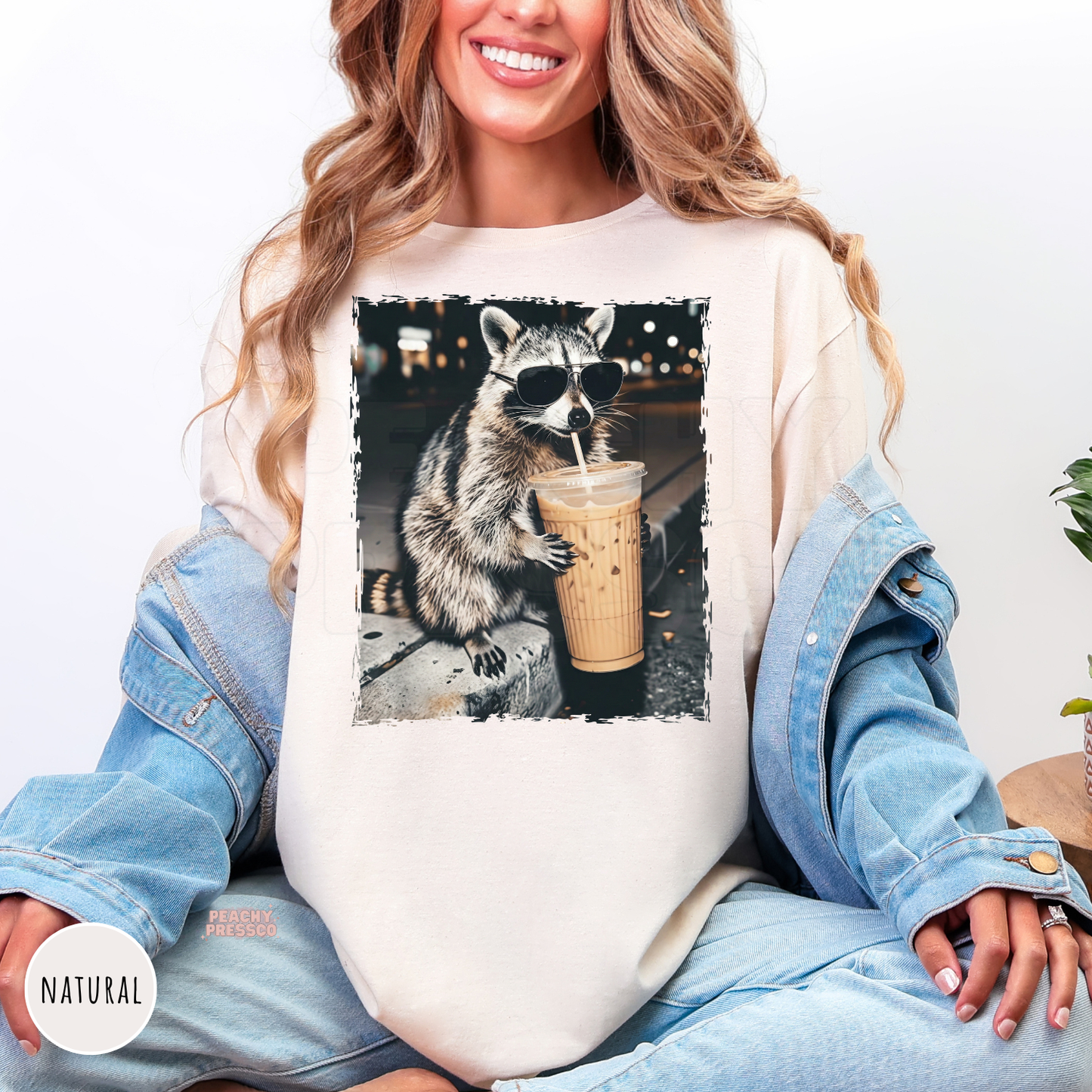 Woman wearing a Natural Gildan graphic t-shirt featuring a funny raccoon wearing sunglasses and sipping an iced coffee. Cute oversized coffee lover shirt with a trendy vintage animal graphic.