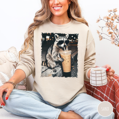 Funny raccoon coffee lover crewneck sweatshirt featuring a sunglasses-wearing raccoon holding an iced coffee on a sand-colored unisex sweatshirt.