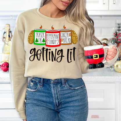A woman wearing a beige crewneck sweatshirt with a Christmas-themed graphic that reads 'Fraser Fir, Peppermint Candy and Gingerbread' and 'Getting Lit' in black script. She is holding a red Santa-themed mug in a decorated kitchen setting.