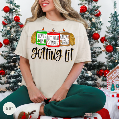 A woman wearing a ivory colored Christmas t-shirt with a holiday-themed graphic that reads 'Getting Lit' with three candles labeled Fraser Fir, Peppermint Candy, and Gingerbread. She is sitting in front of decorated Christmas trees.