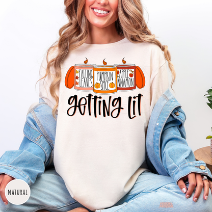 Woman wearing a natural graphic t-shirt featuring a funny "Getting Lit" fall candle design with pumpkin spice, falling leaves, and apple cinnamon artwork.