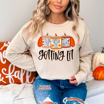 Woman wearing a sand crewneck sweatshirt with a cute "Getting Lit" autumn candle graphic featuring pumpkin spice, falling leaves, and apple cinnamon scents.