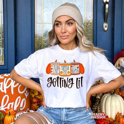 Woman wearing a white t-shirt featuring a funny "Getting Lit" fall graphic with pumpkin spice, falling leaves, and apple cinnamon candles for autumn.