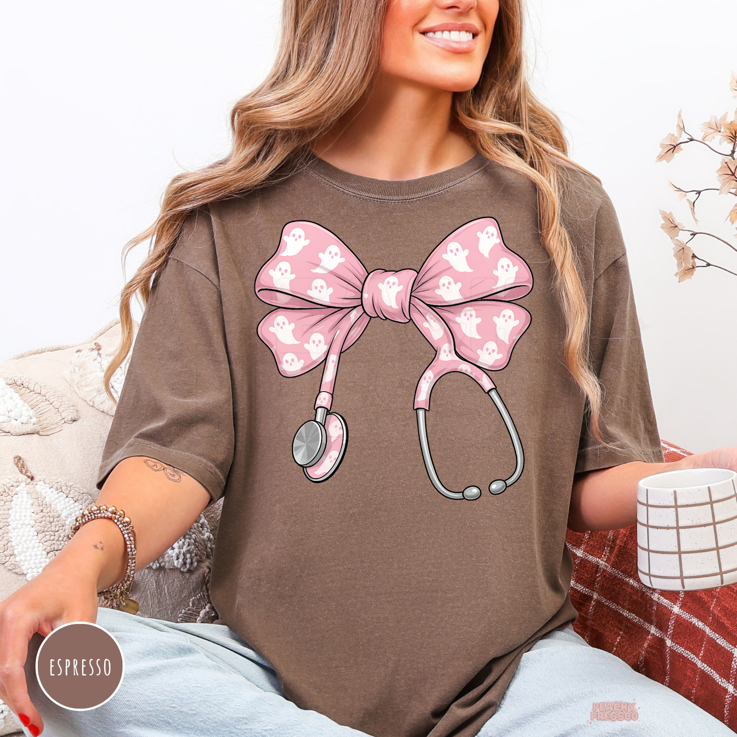 Woman wearing an espresso brown t-shirt featuring a pink ghost stethoscope bow graphic for nurses, doctors, CNAs, and spooky season.