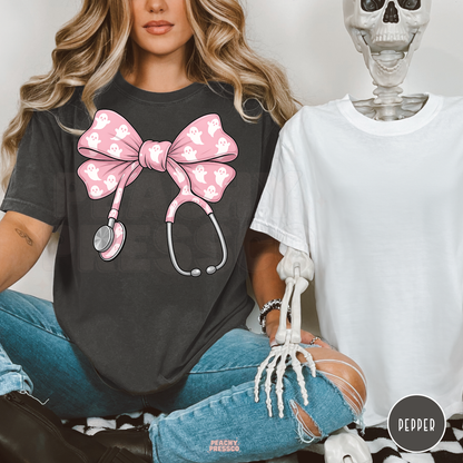 Woman wearing a pepper Comfort Colors t-shirt with a pink ghost stethoscope bow design beside a Halloween skeleton, creating a fun spooky nurse aesthetic.
