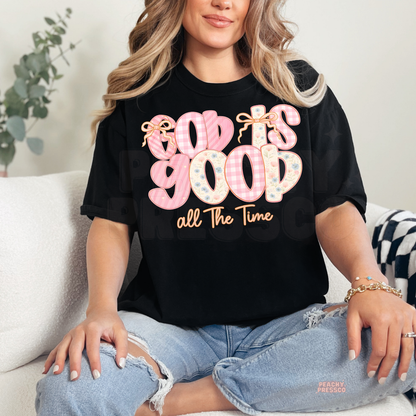 Black unisex t-shirt featuring a pastel "God Is Good All The Time" graphic with floral letters, pink gingham pattern, and bow accents. Christian faith graphic tee with a modern feminine style.