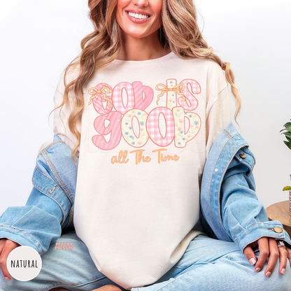 Natural color t-shirt featuring a pastel "God Is Good All The Time" graphic with pink gingham, floral lettering, and delicate bow accents. Soft unisex Christian faith tee with a feminine cottagecore aesthetic.