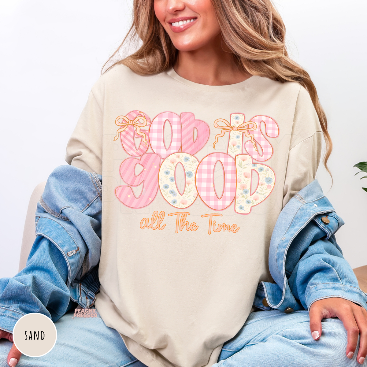 Sand shirt featuring a pastel "God Is Good All The Time" graphic with pink gingham, floral lettering, and bow accents. Soft unisex Christian sweatshirt with a feminine cottagecore look.