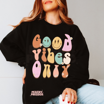 Black sweatshirt featuring a colorful "Good Vibes Only" retro graphic with pastel groovy lettering, smiley faces, daisy flowers, and a fun vintage look.