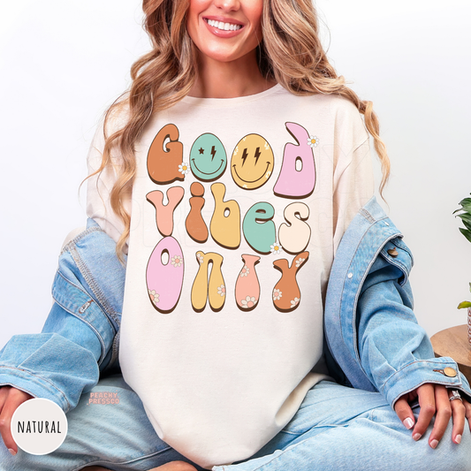 Natural shirt featuring a colorful "Good Vibes Only" graphic with retro pastel lettering, smiley faces, daisy flowers, and a positive boho-inspired design.