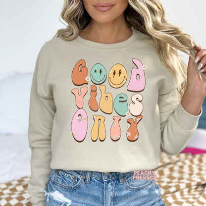 Sand sweatshirt featuring a retro "Good Vibes Only" design with pastel bubble lettering, smiley faces, daisy flowers, and a fun vintage-inspired graphic.