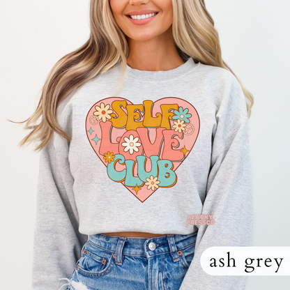 Ash grey sweatshirt showcasing a vintage Self Love Club design with pastel flowers, retro lettering, and a large heart graphic.