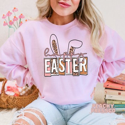 Pink crewneck sweatshirt featuring a Happy Easter leopard bunny ears graphic in soft pastel spring colors.