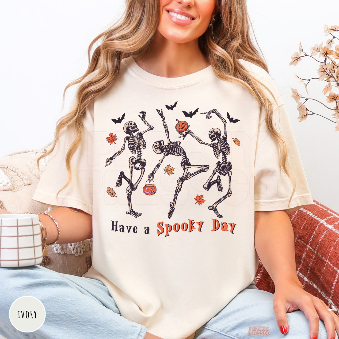 Woman wearing an ivory Comfort Colors shirt featuring vintage dancing skeletons with pumpkins and the phrase "Have a Spooky Day."