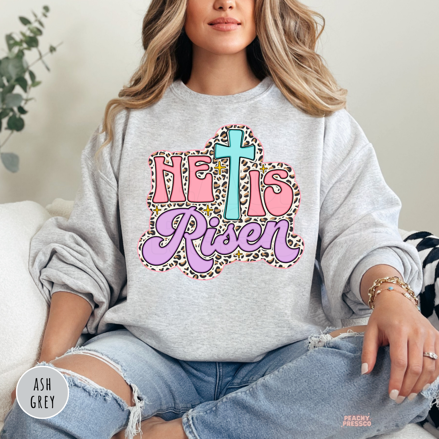 Ash Grey Gildan crewneck sweatshirt featuring a pastel "He Is Risen" design with a turquoise cross, pink lettering, lavender script, leopard print border, and vintage Christian style.