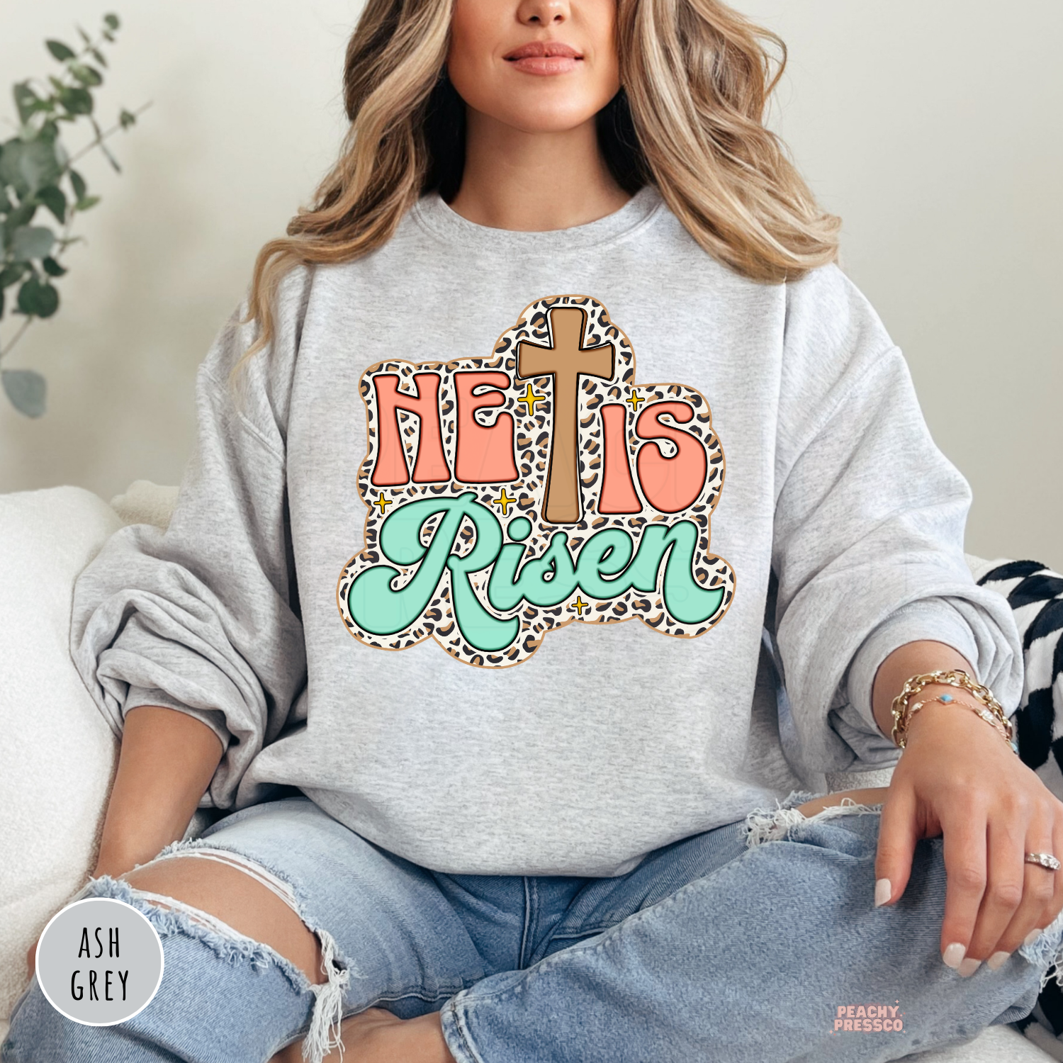 Ash Grey Gildan crewneck sweatshirt featuring a retro "He Is Risen" graphic with a wooden cross, leopard print accents, coral lettering, mint script, and vintage-inspired Christian design.