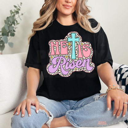 Black unisex t-shirt featuring a pastel "He Is Risen" Christian graphic with a turquoise cross, pink lettering, lavender script, leopard print outline, and sparkling accents. Faith-inspired Easter graphic tee.