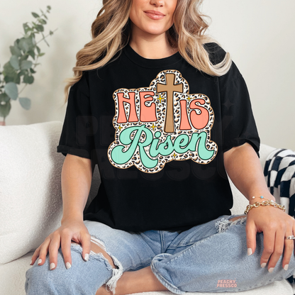 Black Gildan unisex t-shirt featuring a retro "He Is Risen" Christian graphic with a wooden cross, leopard print background, coral lettering, mint script, and sparkling details for Easter and everyday faith wear.