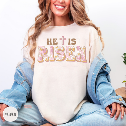 Natural shirt featuring a retro "He Is Risen" floral design with soft pink flowers, vintage brown lettering, and a cross replacing the "T." Christian Easter apparel perfect for celebrating the resurrection.