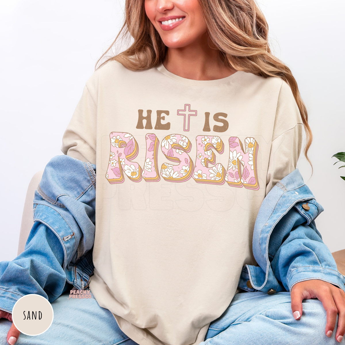 Sand shirt with a vintage "He Is Risen" floral graphic featuring pastel pink floral lettering, brown text, and a cross. Cozy Christian Easter sweatshirt for church, spring, and everyday faith.