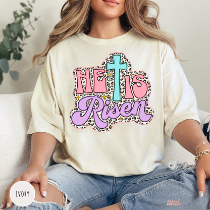 Ivory Comfort Colors garment-dyed t-shirt featuring a pastel "He Is Risen" design with a turquoise cross, pink lettering, lavender script, leopard print border, and sparkling Christian Easter details.