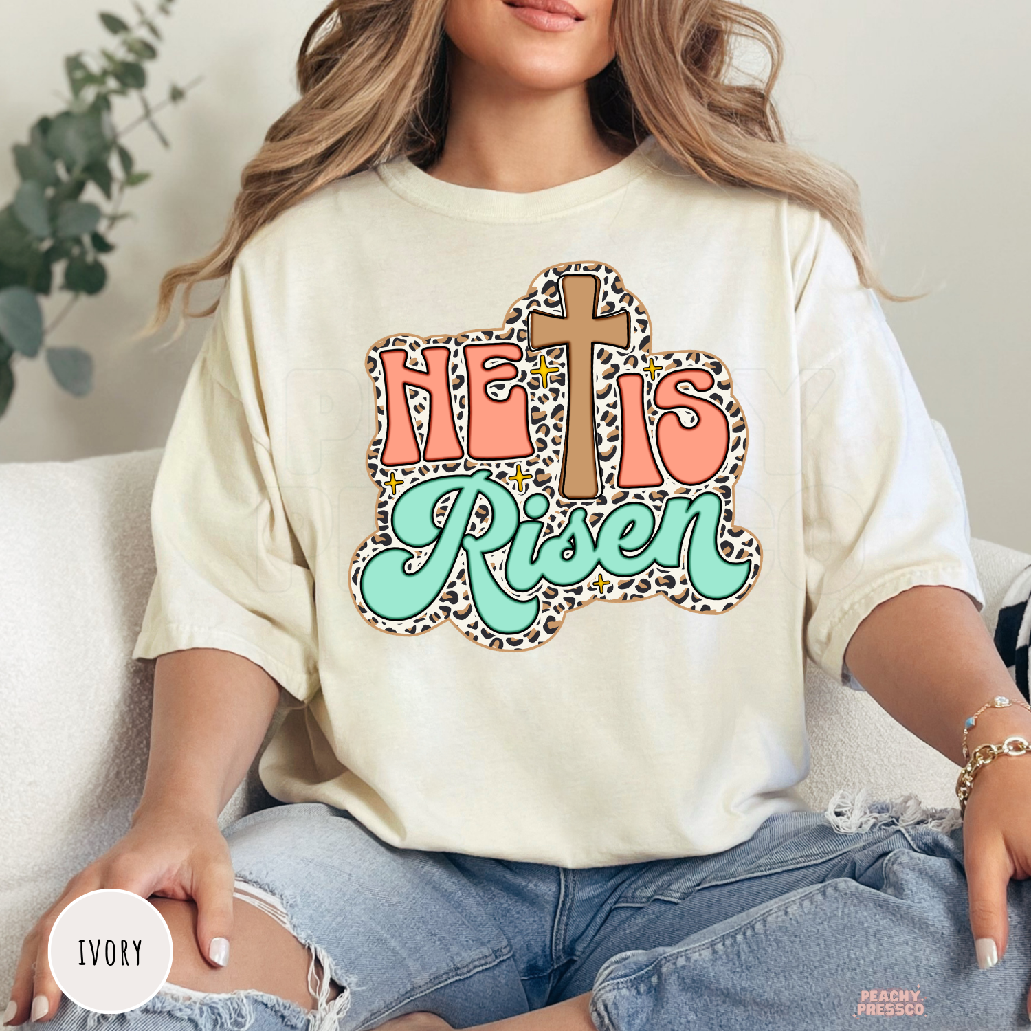 Ivory Comfort Colors garment-dyed t-shirt featuring a retro "He Is Risen" graphic with a wooden cross, leopard print background, coral block lettering, mint script, and gold sparkle details. Relaxed unisex Christian tee.
