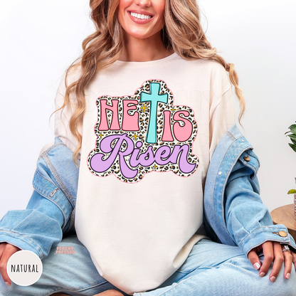 Natural Comfort Colors garment-dyed t-shirt featuring a pastel "He Is Risen" graphic with a turquoise cross, pink lettering, lavender script, leopard print outline, and gold sparkle details. Relaxed unisex Christian tee.