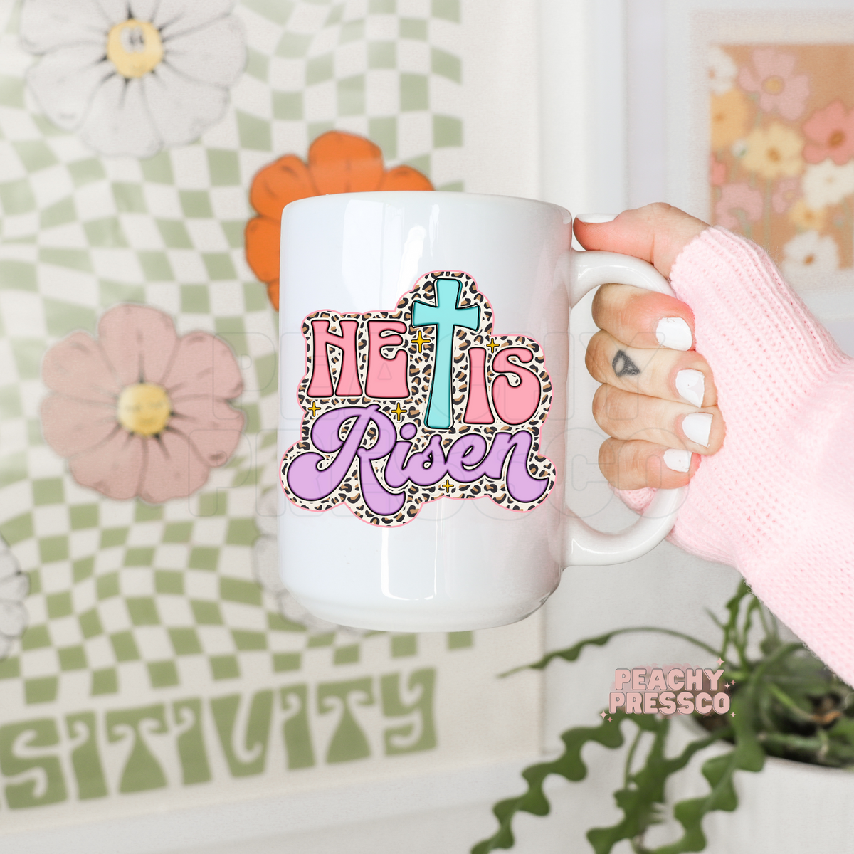 White ceramic coffee mug featuring a pastel "He Is Risen" Christian design with a turquoise cross, pink lettering, lavender script, leopard print border, and sparkling accents. Cute Easter faith coffee mug.
