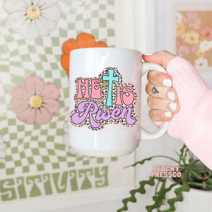 White ceramic coffee mug featuring a pastel "He Is Risen" Christian design with a turquoise cross, pink lettering, lavender script, leopard print border, and sparkling accents. Cute Easter faith coffee mug.