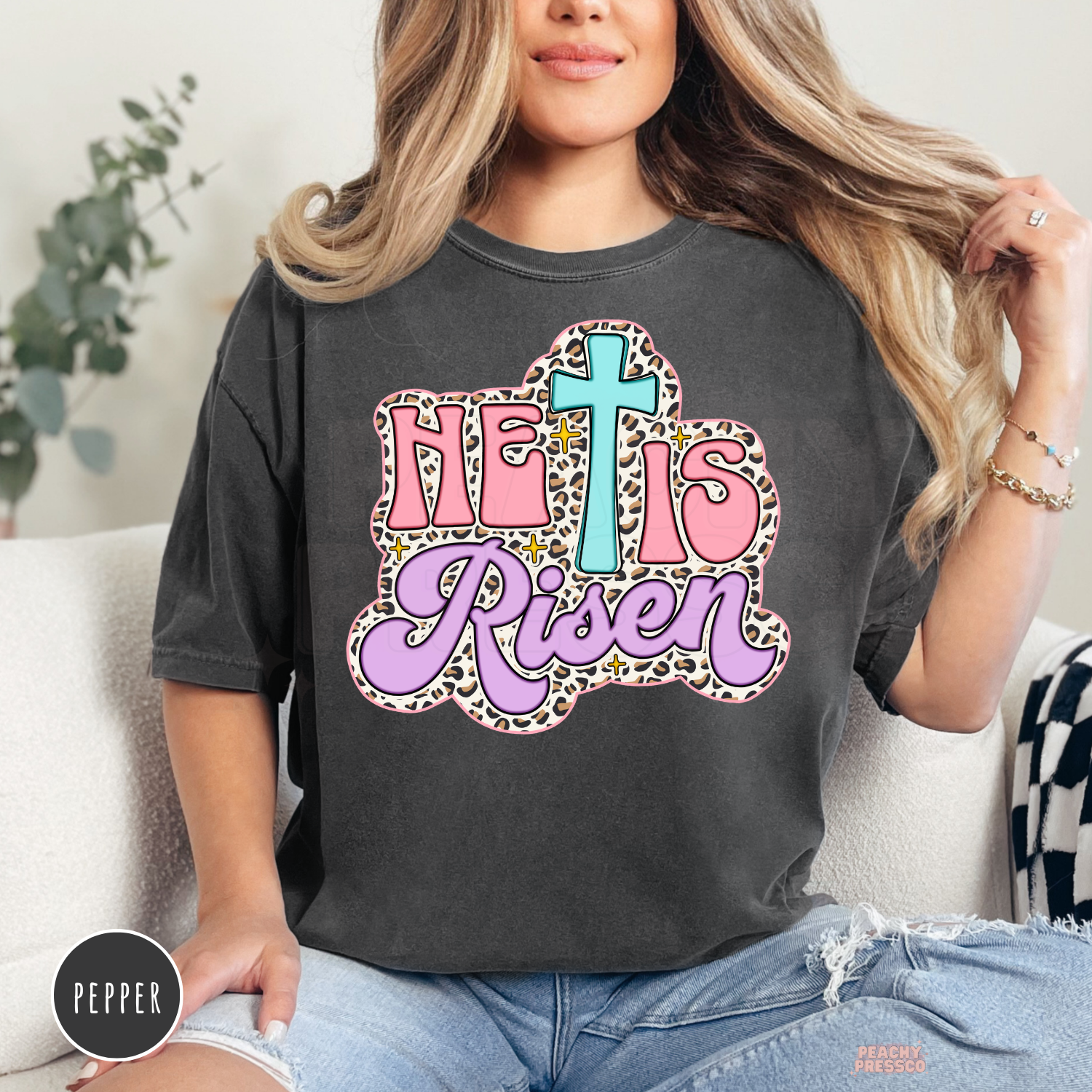 Pepper Comfort Colors garment-dyed t-shirt featuring a pastel "He Is Risen" Christian graphic with a turquoise cross, pink lettering, lavender script, leopard print outline, and retro Easter aesthetic.
