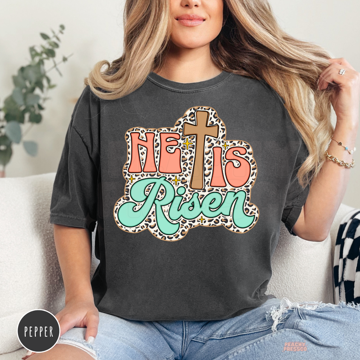 Pepper Comfort Colors garment-dyed t-shirt featuring a retro "He Is Risen" design with a wooden cross, leopard print border, coral lettering, mint green script, and vintage Christian aesthetic.