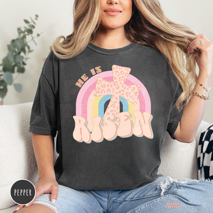 Pepper Comfort Colors t-shirt with a vintage "He Is Risen" design featuring a leopard-print cross, pastel rainbow, and retro lettering. Faith-inspired Christian apparel for Easter.