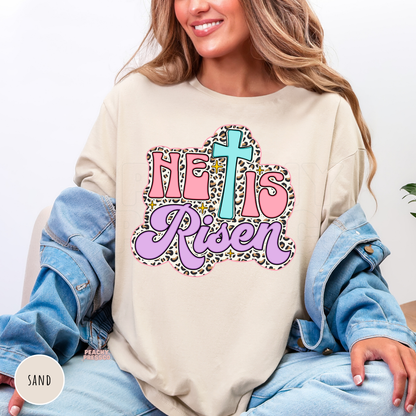 Sand Gildan crewneck sweatshirt featuring a pastel "He Is Risen" Christian graphic with a turquoise cross, pink lettering, lavender script, leopard print accents, and vintage-inspired Easter style.