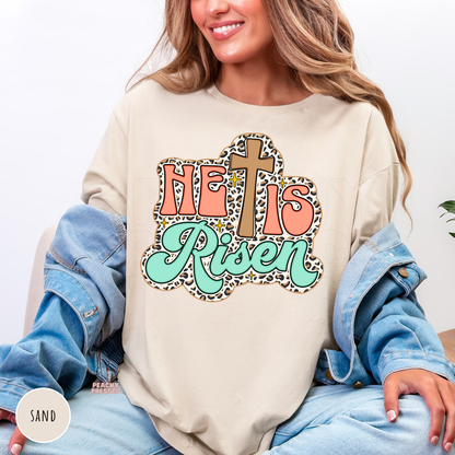 Sand shirt featuring a vintage "He Is Risen" design with a wooden cross, leopard print border, coral lettering, mint script, and subtle sparkle accents. Cozy unisex Christian Easter sweatshirt.