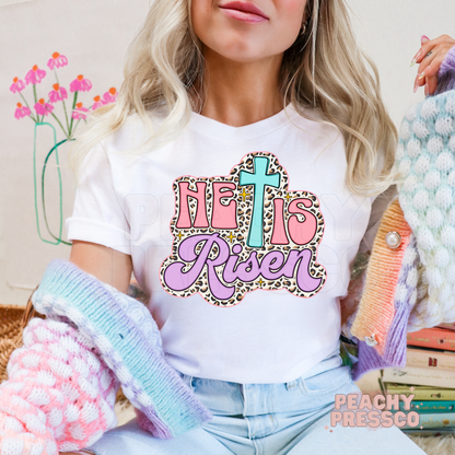 White unisex t-shirt featuring a pastel "He Is Risen" Christian design with a turquoise cross, pink lettering, lavender script, leopard print background, and sparkling accents for Easter.