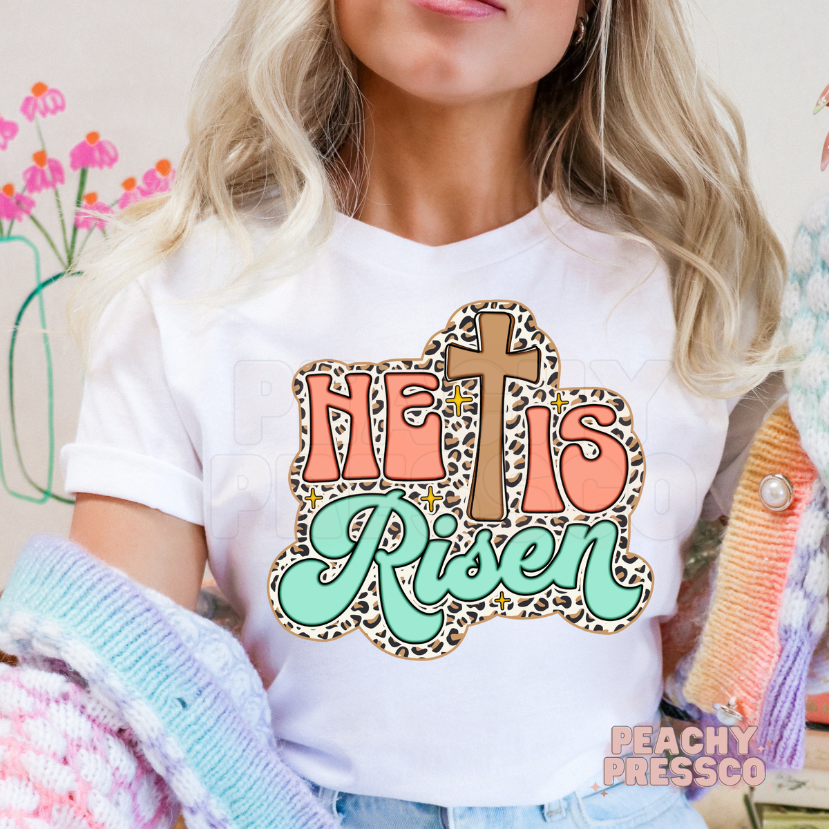 White unisex t-shirt featuring a retro "He Is Risen" Christian design with a wooden cross, leopard print outline, coral lettering, mint script, and sparkling accents. Faith-inspired Easter graphic tee.