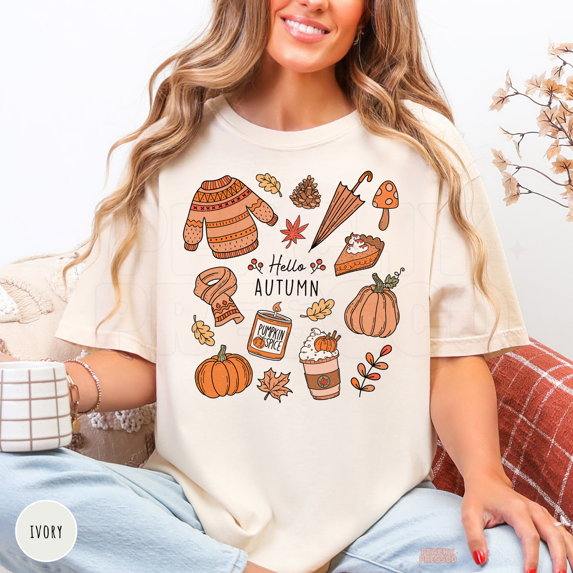 Woman wearing an ivory Comfort Colors t-shirt featuring a Hello Autumn design with pumpkins, pumpkin spice coffee, cozy sweater, scarf, umbrella, pie, and autumn leaves.