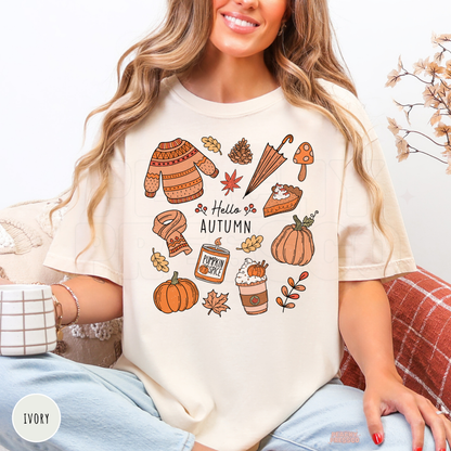 Woman wearing an ivory Comfort Colors t-shirt featuring a Hello Autumn design with pumpkins, pumpkin spice coffee, cozy sweater, scarf, umbrella, pie, and autumn leaves.