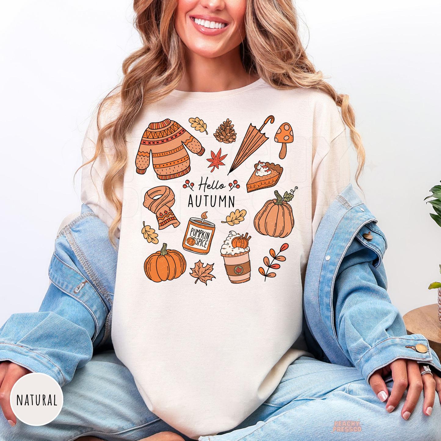 Woman wearing a natural graphic t-shirt featuring a cute Hello Autumn design with pumpkins, pumpkin spice latte, sweater, scarf, umbrella, pie, mushrooms, and cozy fall illustrations.