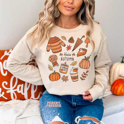 Woman wearing a sand crewneck sweatshirt featuring a Hello Autumn design with pumpkins, pumpkin spice candle, coffee, sweater, scarf, umbrella, pie, and fall leaves.
