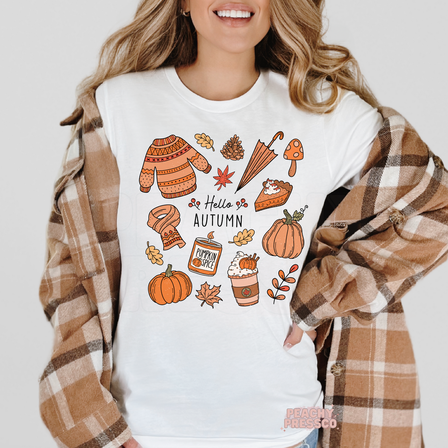 Woman wearing a white t-shirt layered with a plaid flannel featuring a Hello Autumn graphic with pumpkins, pumpkin spice latte, sweater, scarf, mushrooms, pie, and cozy fall elements.