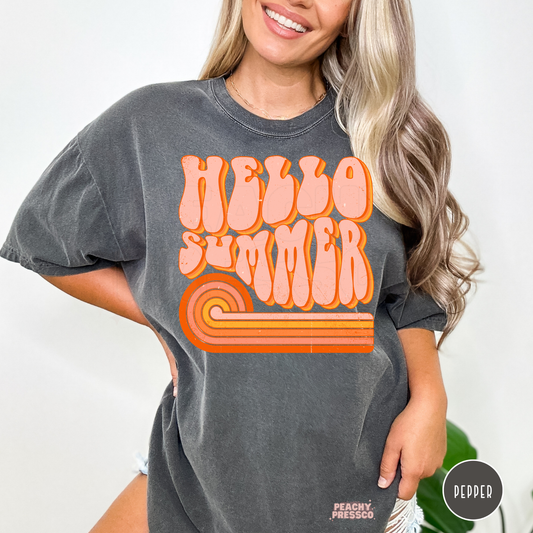 Woman wearing a pepper Comfort Colors t-shirt featuring a retro "Hello Summer" graphic with groovy lettering and a vintage orange rainbow design.