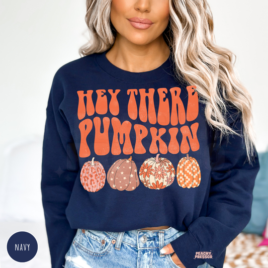 Woman wearing a navy crewneck sweatshirt featuring a retro "Hey There Pumpkin" graphic with four patterned pumpkins in pink, cream, and orange fall colors.