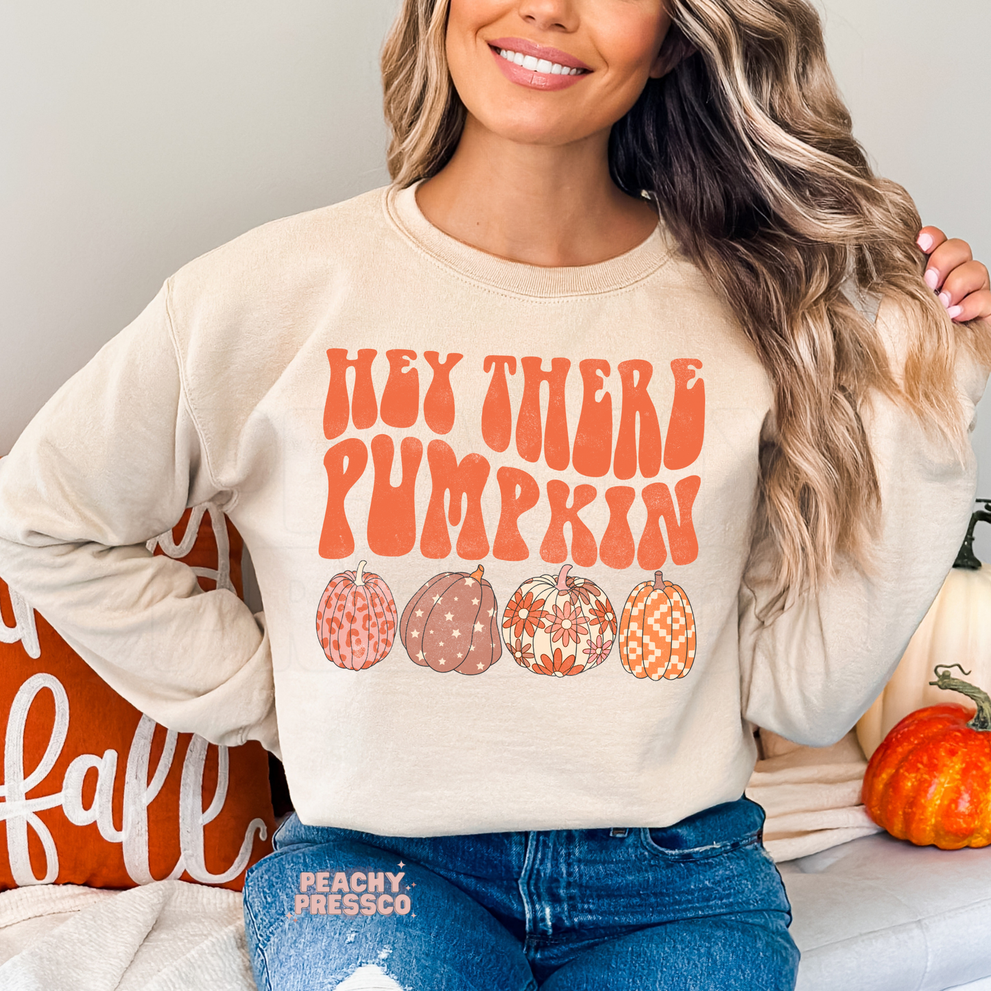 Woman wearing a sand sweatshirt featuring the phrase "Hey There Pumpkin" with four retro patterned pumpkins in warm autumn colors.
