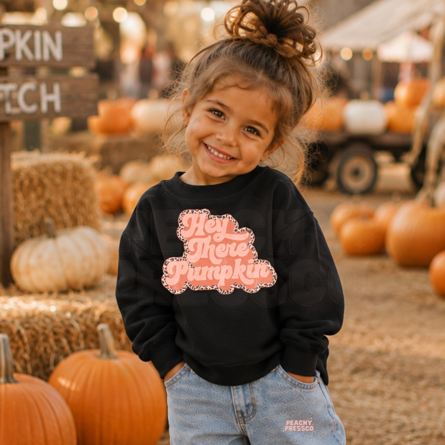 Happy toddler girl wearing a black "Hey There Pumpkin" sweatshirt with a retro pink design at a pumpkin patch surrounded by pumpkins.