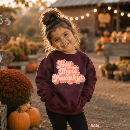 Toddler girl wearing a maroon "Hey There Pumpkin" sweatshirt with a retro pink graphic in a rustic pumpkin patch during autumn.